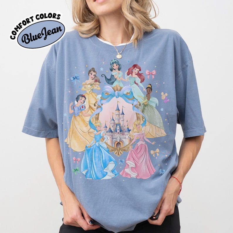 May include: Blue t-shirt with a graphic of Disney princesses surrounding a castle. The shirt has the words "Comfort Colors" and "Blue Jean" printed on it. The princesses are in various gowns, with a light blue background.