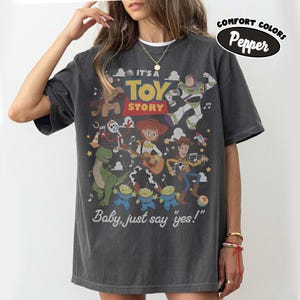 May include: A charcoal gray short-sleeve t-shirt featuring a graphic print of Toy Story characters including Woody, Buzz Lightyear, Jessie, Rex, Forky, and the aliens. The text reads IT'S A TOY STORY and Baby, just say yes! with a Comfort Colors Pepper label.