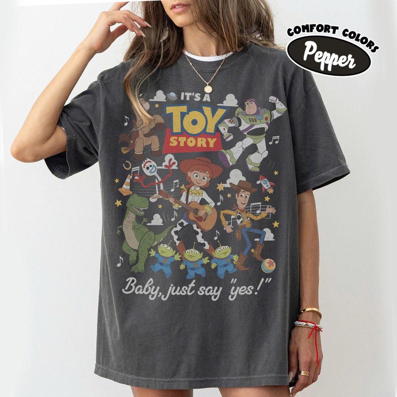May include: A charcoal gray short-sleeve t-shirt featuring a graphic print of Toy Story characters including Woody, Buzz Lightyear, Jessie, Rex, Forky, and the aliens. The text reads IT'S A TOY STORY and Baby, just say yes! with a Comfort Colors Pepper label.