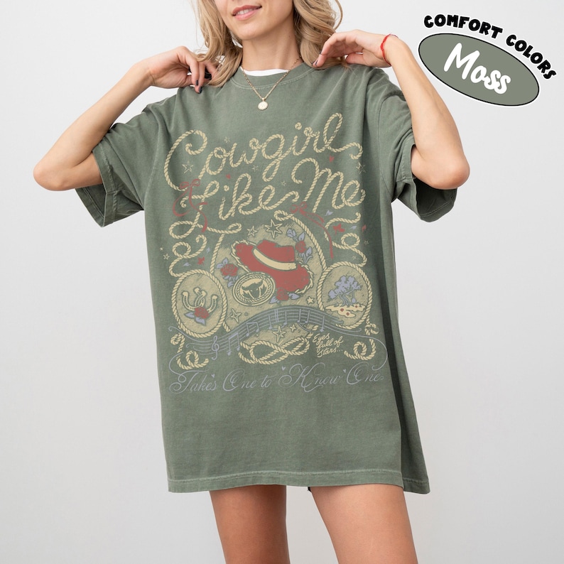 May include: A moss green short-sleeve t-shirt featuring a graphic print with rope-style lettering that reads Cowgirl Like Me and Takes One to Know One. The design includes a red cowboy hat, a horseshoe, and musical notes in a western style.