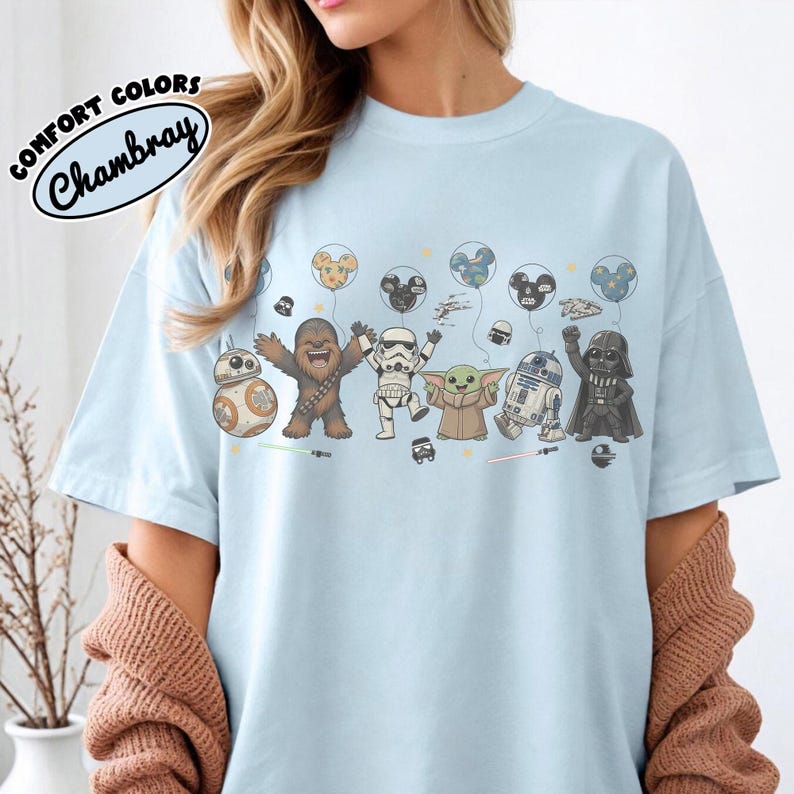 K&ouml;nnte beinhalten: Ein hellblaues T-Shirt mit einem Grafikdruck verschiedener Star Wars-Figuren, darunter Chewbacca, ein Stormtrooper, Grogu, R2-D2 und Darth Vader, sowie schwebende Luftballons in Form von Mickey-Maus-Ohren und kleinen Raumschiffen.