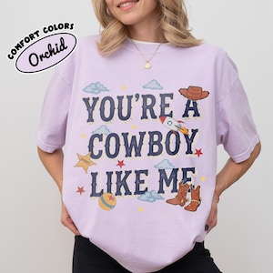May include: A light purple t-shirt featuring the text "YOU'RE A COWBOY LIKE ME" in dark blue letters. The design includes illustrations of a brown cowboy hat, a rocket, a yellow sheriff star, a ball, and brown cowboy boots, with small clouds and stars.