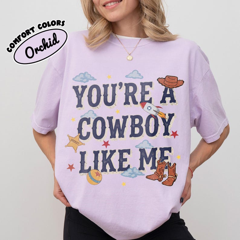 May include: A light purple t-shirt featuring the text "YOU'RE A COWBOY LIKE ME" in dark blue letters. The design includes illustrations of a brown cowboy hat, a rocket, a yellow sheriff star, a ball, and brown cowboy boots, with small clouds and stars.
