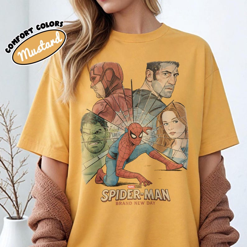 May include: Mustard-colored t-shirt featuring a graphic of Spider-Man and other Marvel characters. The design includes the text "SPIDER-MAN BRAND NEW DAY". The shirt has short sleeves and a crew neck.