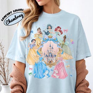 May include: Light blue t-shirt featuring a colorful illustration of several princesses surrounding a castle. The shirt has the words "COMFORT COLORS" and "Chambray" printed on the upper left side.
