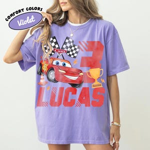 Puede incluir: Camiseta violeta Comfort Colors con un coche de carreras de dibujos animados rojo, banderas a cuadros, un trofeo dorado y el nombre "Lucas" en letras rojas. El n&uacute;mero "3" tambi&eacute;n est&aacute; impreso en la camiseta.