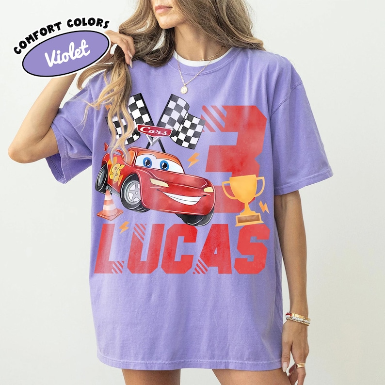 Puede incluir: Camiseta violeta Comfort Colors con un coche de carreras de dibujos animados rojo, banderas a cuadros, un trofeo dorado y el nombre "Lucas" en letras rojas. El n&uacute;mero "3" tambi&eacute;n est&aacute; impreso en la camiseta.