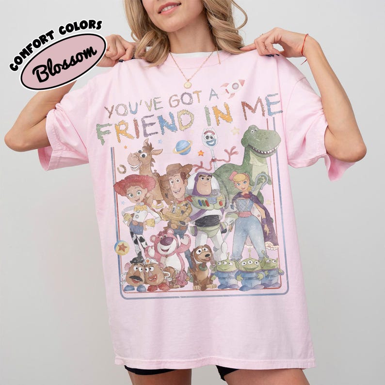 May include: A light pink t-shirt featuring a graphic print of various animated toy characters. The text "YOU'VE GOT A FRIEND IN ME" is printed above the characters in a colorful, distressed font. A small oval logo reads "COMFORT COLORS Blossom".