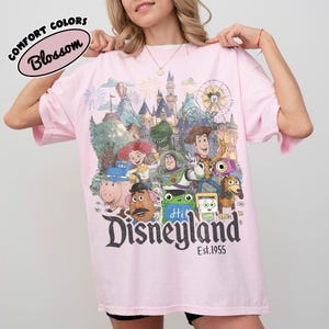 May include: A light pink t-shirt featuring a graphic print of various animated characters, a castle, and a Ferris wheel. The text reads "Disneyland Est. 1955" at the bottom, with a "Comfort Colors Blossom" label in the upper left corner of the image.