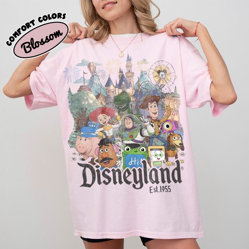 May include: A light pink t-shirt featuring a graphic print of various animated characters, a castle, and a Ferris wheel. The text reads "Disneyland Est. 1955" at the bottom, with a "Comfort Colors Blossom" label in the upper left corner of the image.