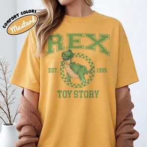 Pu&ograve; includere: T-shirt color senape con un dinosauro verde dei cartoni animati, la parola "REX" e il testo "TOY STORY". La maglietta include anche il testo "EST. 1995" in verde. La maglietta &egrave; in stile comfort colors.