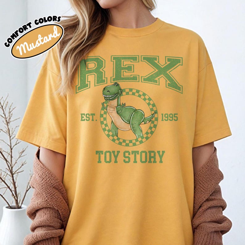 Pu&ograve; includere: T-shirt color senape con un dinosauro verde dei cartoni animati, la parola "REX" e il testo "TOY STORY". La maglietta include anche il testo "EST. 1995" in verde. La maglietta &egrave; in stile comfort colors.