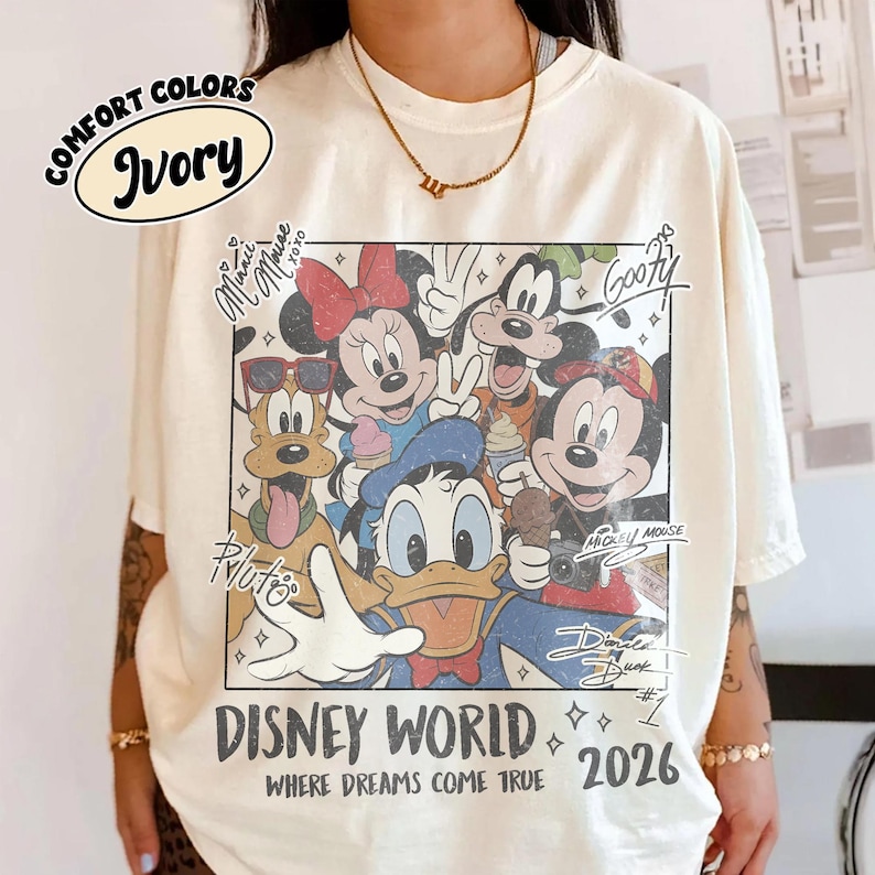 May include: Ivory-colored t-shirt featuring a Disney World graphic with Mickey Mouse, Minnie Mouse, Donald Duck, Goofy, and Pluto. The graphic includes the text "DISNEY WORLD WHERE DREAMS COME TRUE 2026". The shirt has short sleeves and a crew neck.