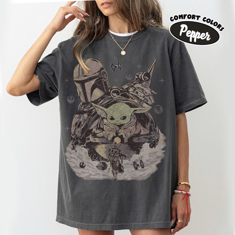 Puede incluir: Camiseta gris oscuro con un gr&aacute;fico de The Mandalorian, Grogu y una nave espacial. El dise&ntilde;o incluye el texto "Comfort Colors Pepper" en un &oacute;valo negro. La camiseta tiene mangas cortas y un corte holgado.