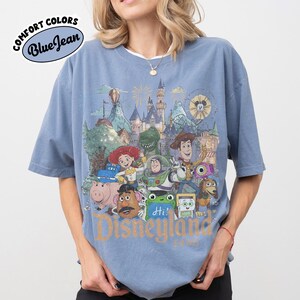 May include: A blue short-sleeve t-shirt featuring a graphic print of various animated characters in front of a castle. The text "Disneyland" is printed in gold at the bottom. A "Comfort Colors Blue Jean" logo is visible in the upper left corner.
