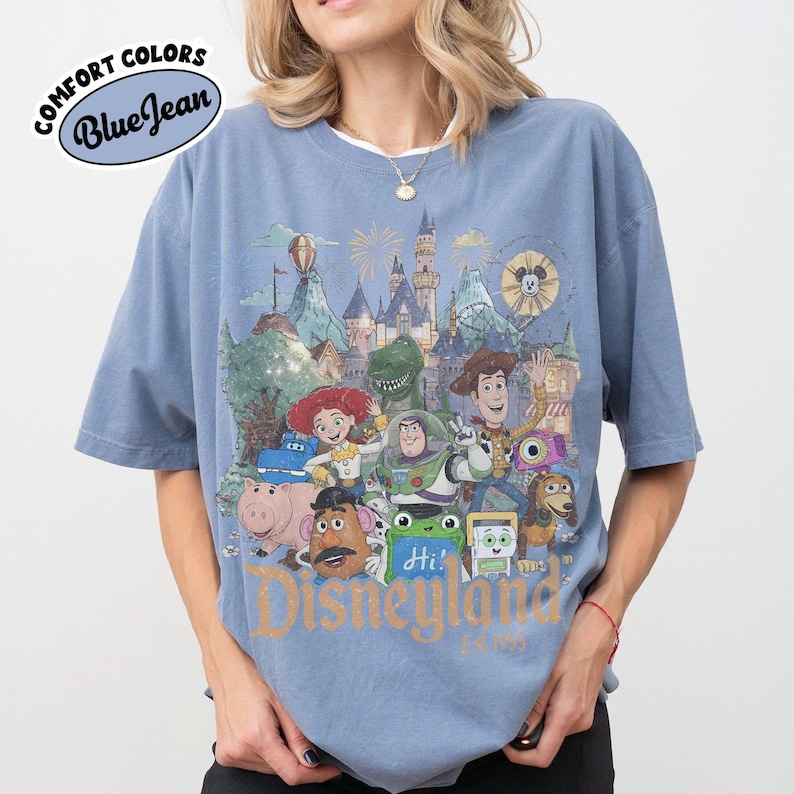 May include: A blue short-sleeve t-shirt featuring a graphic print of various animated characters in front of a castle. The text "Disneyland" is printed in gold at the bottom. A "Comfort Colors Blue Jean" logo is visible in the upper left corner.