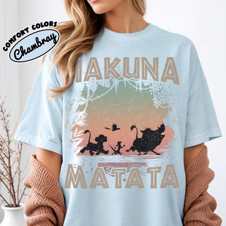 May include: Pale blue t-shirt with the words "Hakuna Matata" in tan lettering. The shirt features a silhouette of Simba, Timon, Pumbaa, and a bird. The shirt has the words "Comfort Colors Chambray" in a circle.