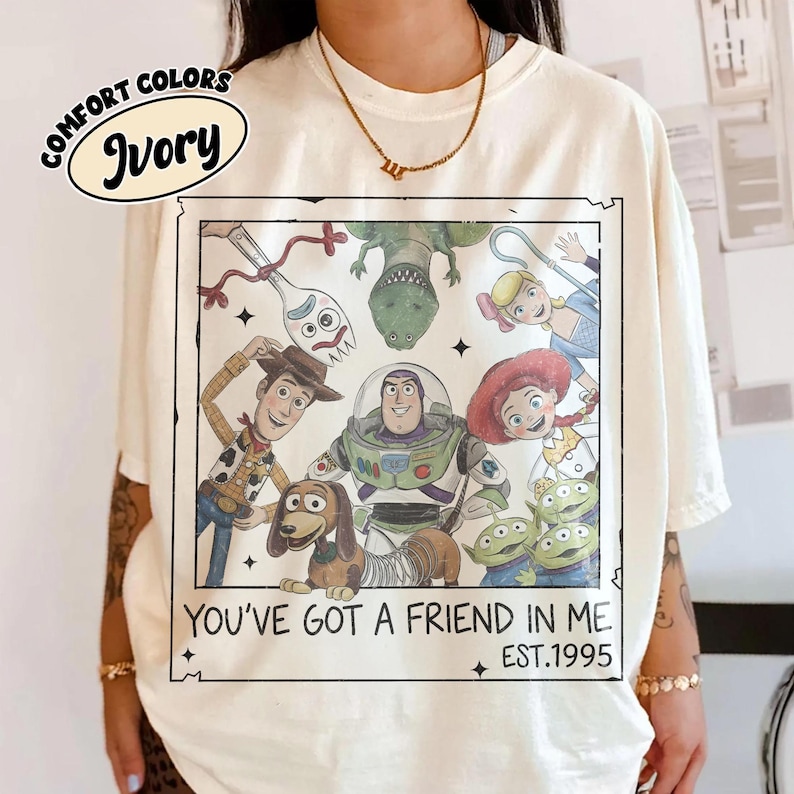 May include: Ivory-colored t-shirt featuring a graphic of Toy Story characters. The graphic includes Woody, Buzz Lightyear, and other characters from the movie. Text on the shirt reads "YOU'VE GOT A FRIEND IN ME" and "EST.1995".