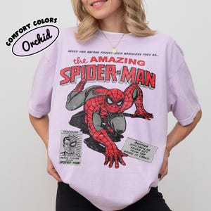 May include: A light purple short-sleeve t-shirt featuring a distressed graphic of Spider-Man in red and black. The text reads: COMFORT COLORS Orchid, NEVER HAS ANYONE FOUGHT SUCH MERCILESS FOES AS... the AMAZING SPIDER-MAN, FEATURING PETER PARKER, and more.