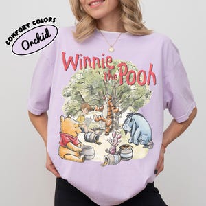 May include: A light purple t-shirt featuring a graphic of Winnie the Pooh and friends. The design includes Winnie the Pooh, Tigger, Piglet, Eeyore, and Christopher Robin. The shirt has the words "Winnie the Pooh" in red script.