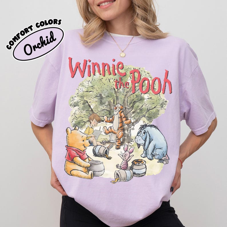 May include: A light purple t-shirt featuring a graphic of Winnie the Pooh and friends. The design includes Winnie the Pooh, Tigger, Piglet, Eeyore, and Christopher Robin. The shirt has the words "Winnie the Pooh" in red script.