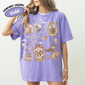 May include: Violet Comfort Colors t-shirt featuring cartoon characters in ornate frames and decorative elements. The shirt has a relaxed fit and a crew neck. The text "Comfort Colors" and "Violet" are in a bubble on the top left.