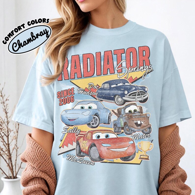 May include: Light blue t-shirt featuring a graphic of characters from the movie Cars, including Doc Hudson, Sally, Mater, and Lightning McQueen. The design includes the text "Radiator Springs" and "Since 2006". The shirt has a vintage, retro style.