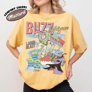 May include: Mustard-coloured t-shirt featuring a graphic of Buzz Lightyear, aliens, and Zurg from the movie Toy Story. The shirt has the words "BUZZ lightyear" and "LASER BLAST" printed on it. The shirt is a comfort colours style.