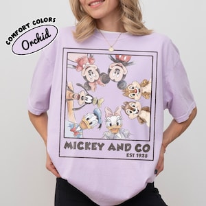 Puede incluir: Camiseta color orqu&iacute;dea con un gr&aacute;fico enmarcado de Mickey Mouse, Minnie Mouse, Donald Duck, Daisy Duck, Goofy y Chip y Chop. El texto "Mickey and Co. Est. 1928" est&aacute; impreso debajo. La camiseta es de la marca Comfort Colors.