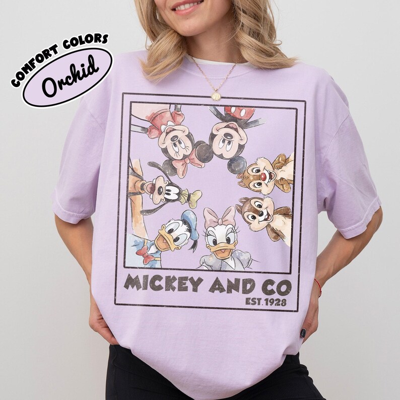 Puede incluir: Camiseta color orqu&iacute;dea con un gr&aacute;fico enmarcado de Mickey Mouse, Minnie Mouse, Donald Duck, Daisy Duck, Goofy y Chip y Chop. El texto "Mickey and Co. Est. 1928" est&aacute; impreso debajo. La camiseta es de la marca Comfort Colors.