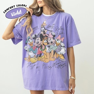 May include: Violet Comfort Colors t-shirt featuring a graphic of Disney characters including Mickey Mouse, Minnie Mouse, Donald Duck, Daisy Duck, Goofy, and Pluto. The shirt has signatures and the text "MOUSE" and "xoxo".