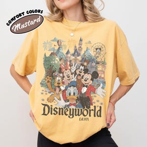 May include: Mustard-colored t-shirt featuring a graphic of Disney characters in front of a castle, fireworks, and a Ferris wheel. The text "Disneyworld Est.1971" is printed below the graphic. The shirt has short sleeves and a crew neck.