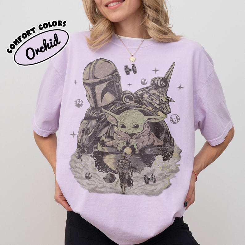 Puede incluir: Una camiseta de color lila claro con un gr&aacute;fico de personajes de una serie de ciencia ficci&oacute;n. El gr&aacute;fico incluye una figura con casco, una peque&ntilde;a criatura verde y una nave espacial. El texto "COMFORT COLORS Orchid" est&aacute; impreso en la parte superior izquierda.
