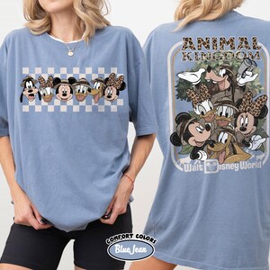 K&ouml;nnte beinhalten: Ein blaues Comfort Colors T-Shirt mit einer Cartoon-Grafik von Disney-Figuren. Die Vorderseite zeigt eine Reihe von Figuren auf kariertem Hintergrund. Die R&uuml;ckseite hat eine gr&ouml;&szlig;ere Grafik mit dem Text "ANIMAL KINGDOM" und "Walt Disney World".
