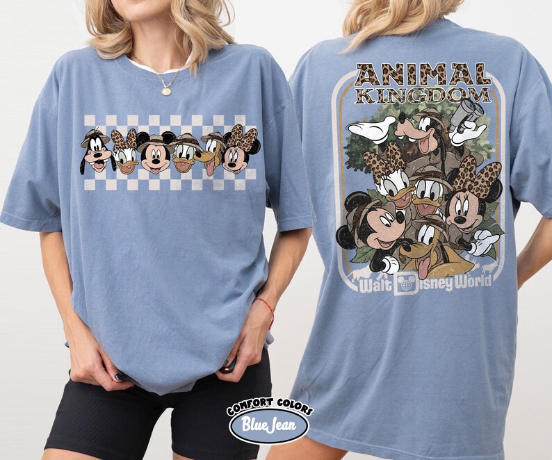 K&ouml;nnte beinhalten: Ein blaues Comfort Colors T-Shirt mit einer Cartoon-Grafik von Disney-Figuren. Die Vorderseite zeigt eine Reihe von Figuren auf kariertem Hintergrund. Die R&uuml;ckseite hat eine gr&ouml;&szlig;ere Grafik mit dem Text "ANIMAL KINGDOM" und "Walt Disney World".