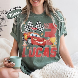 Puede incluir: Camiseta verde con un dise&ntilde;o tem&aacute;tico de Cars. El dise&ntilde;o incluye un coche de carreras rojo, banderas a cuadros, un trofeo dorado, el n&uacute;mero 3 y el nombre "LUCAS" en rojo. La camiseta tambi&eacute;n tiene el texto "COMFORT COLORS" y "Moss".