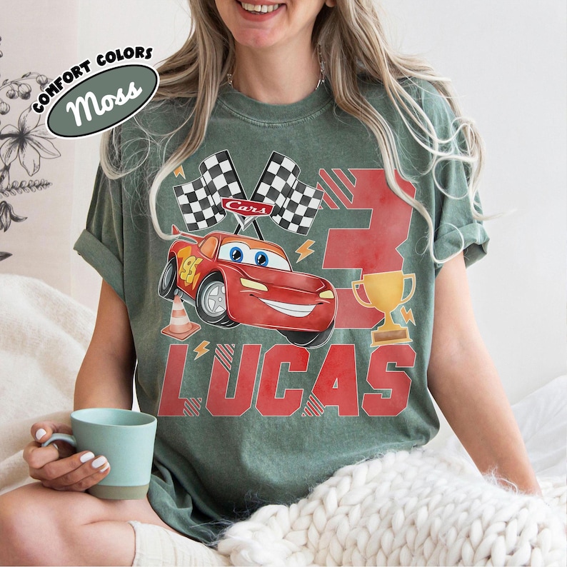 Puede incluir: Camiseta verde con un dise&ntilde;o tem&aacute;tico de Cars. El dise&ntilde;o incluye un coche de carreras rojo, banderas a cuadros, un trofeo dorado, el n&uacute;mero 3 y el nombre "LUCAS" en rojo. La camiseta tambi&eacute;n tiene el texto "COMFORT COLORS" y "Moss".