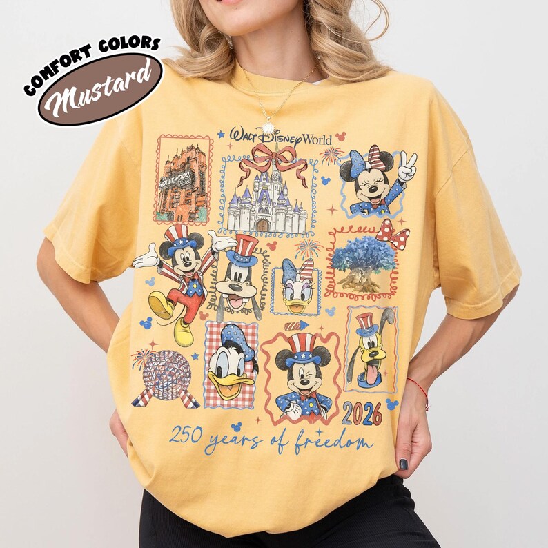 May include: Mustard-colored t-shirt featuring a collage of Disney characters and landmarks, including Mickey Mouse, Minnie Mouse, and Cinderella's castle. The shirt has the text "Walt Disney World" and "250 years of freedom 2026".