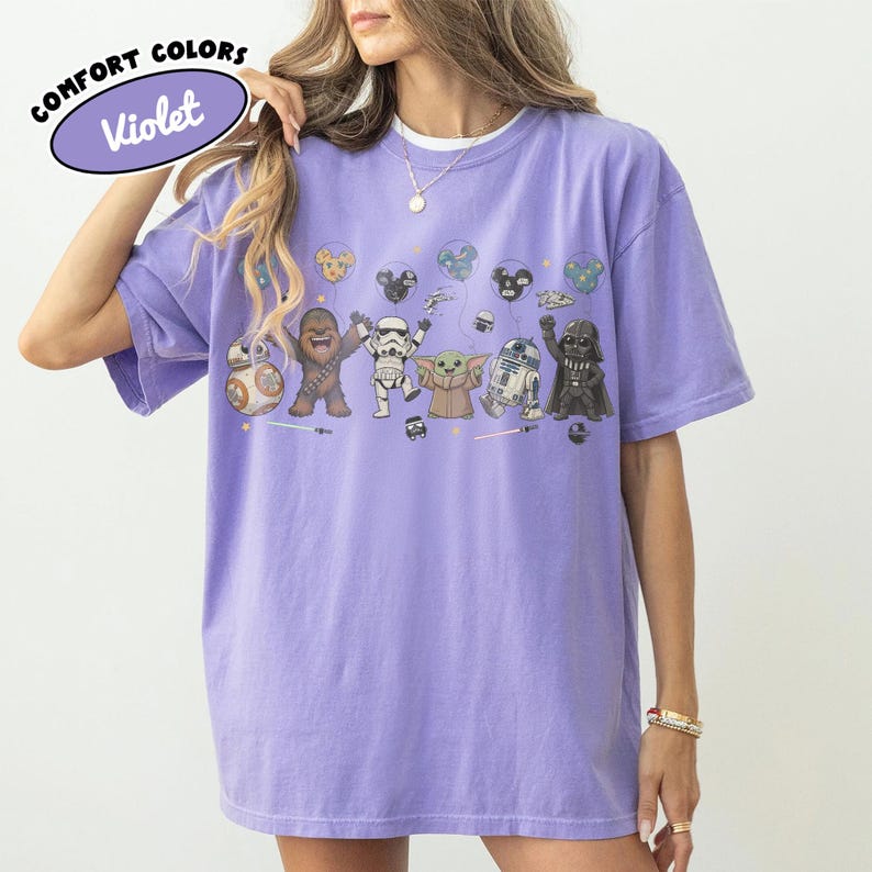 K&ouml;nnte beinhalten: Ein violettes kurz&auml;rmeliges T-Shirt mit einem Grafikdruck verschiedener Star Wars-Figuren, darunter Chewbacca, ein Sturmtruppler, Grogu, R2-D2 und Darth Vader, sowie schwebenden Luftballons und kleinen Symbolen. Der Text Comfort Colors Violet ist sichtbar.