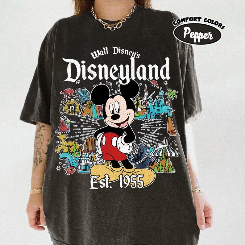 Op de afbeelding: Een antracietgrijs t-shirt met korte mouwen en een afbeelding van Mickey Mouse boven de tekst Est. 1955. Het ontwerp bevat kleurrijke illustraties van Disneyland-attracties en de tekst Walt Disney's Disneyland. Comfort Colors Pepper.
