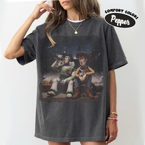 May include: A charcoal grey short-sleeve t-shirt featuring a graphic print of Buzz Lightyear and Woody sitting together at night. The shirt has a crew neckline and a relaxed fit. A circular label in the top right corner reads Comfort Colors Pepper.