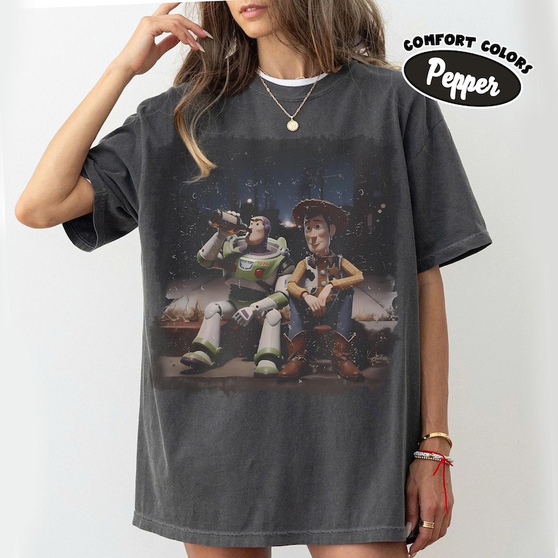 May include: A charcoal grey short-sleeve t-shirt featuring a graphic print of Buzz Lightyear and Woody sitting together at night. The shirt has a crew neckline and a relaxed fit. A circular label in the top right corner reads Comfort Colors Pepper.