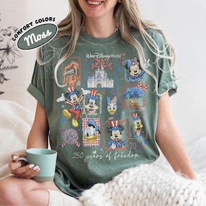 May include: Moss green t-shirt with a patriotic Disney World design. The design includes Mickey Mouse, Minnie Mouse, Donald Duck, and Goofy in patriotic attire, along with the text "250 years of freedom" and "2026".
