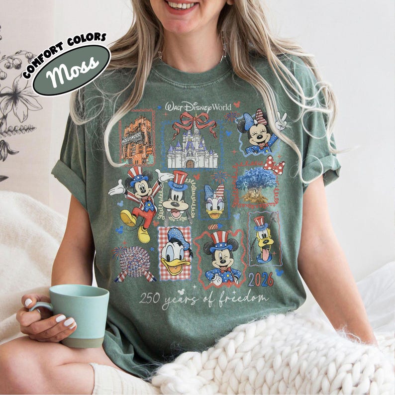 May include: Moss green t-shirt with a patriotic Disney World design. The design includes Mickey Mouse, Minnie Mouse, Donald Duck, and Goofy in patriotic attire, along with the text "250 years of freedom" and "2026".