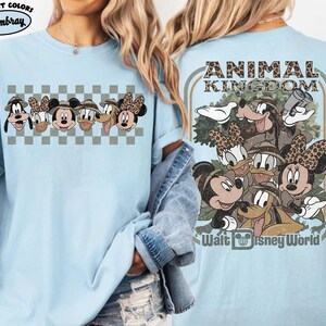 K&ouml;nnte beinhalten: Hellblaues T-Shirt mit einem Disney Animal Kingdom-Design. Auf der Vorderseite sind Gesichter von Charakteren zu sehen, w&auml;hrend die R&uuml;ckseite eine Gruppe von Charakteren mit dem Text "Animal Kingdom" und "Walt Disney World" zeigt.