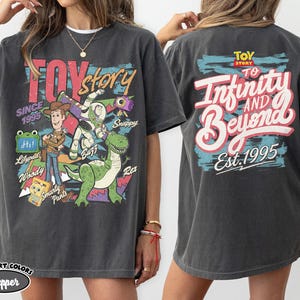 Pode incluir: Uma camiseta de manga curta cinza-carv&atilde;o com gr&aacute;ficos de Toy Story. A frente exibe Woody, Buzz Lightyear, Rex e texto incluindo "Toy Story" e "Since 1995". As costas apresentam o texto "Toy Story To Infinity and Beyond Est. 1995".