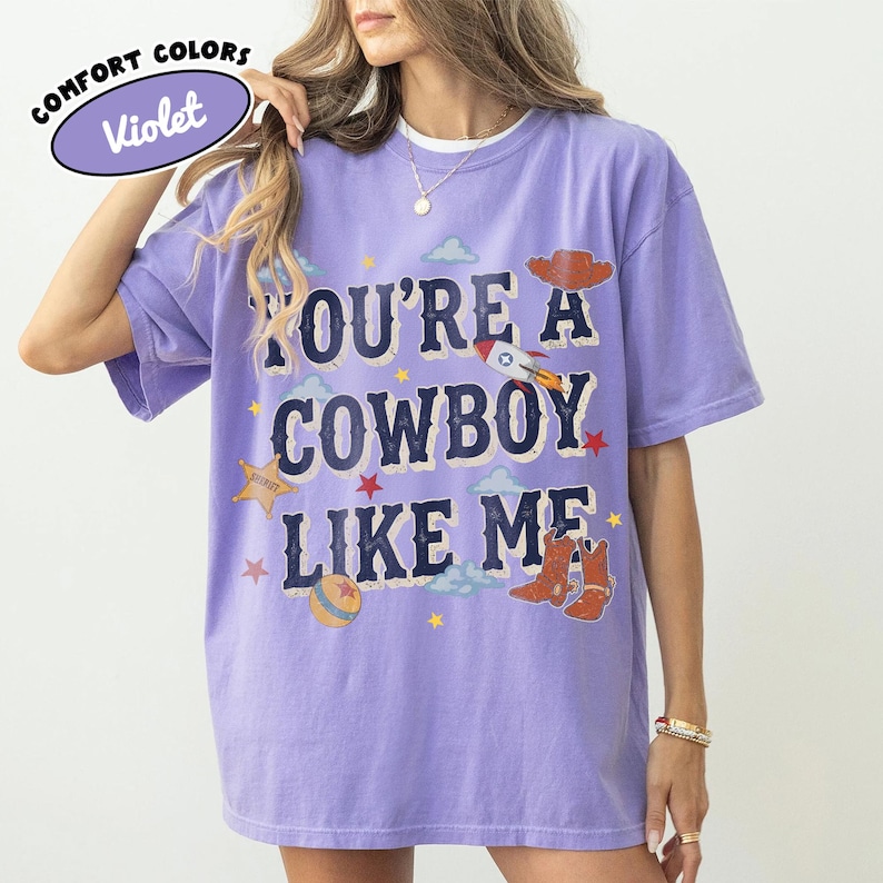 May include: A violet short-sleeve t-shirt featuring the text "YOU'RE A COWBOY LIKE ME" in dark blue letters. The graphic includes a brown cowboy hat, a rocket ship, a sheriff star, a ball, and brown cowboy boots, accented by small stars and clouds.