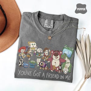 May include: A charcoal gray crew neck t-shirt featuring a colorful graphic print of various animated toy characters. Below the characters, the text reads, "YOU'VE GOT A FRIEND IN ME." The shirt has a Comfort Colors brand label at the inner neckline.