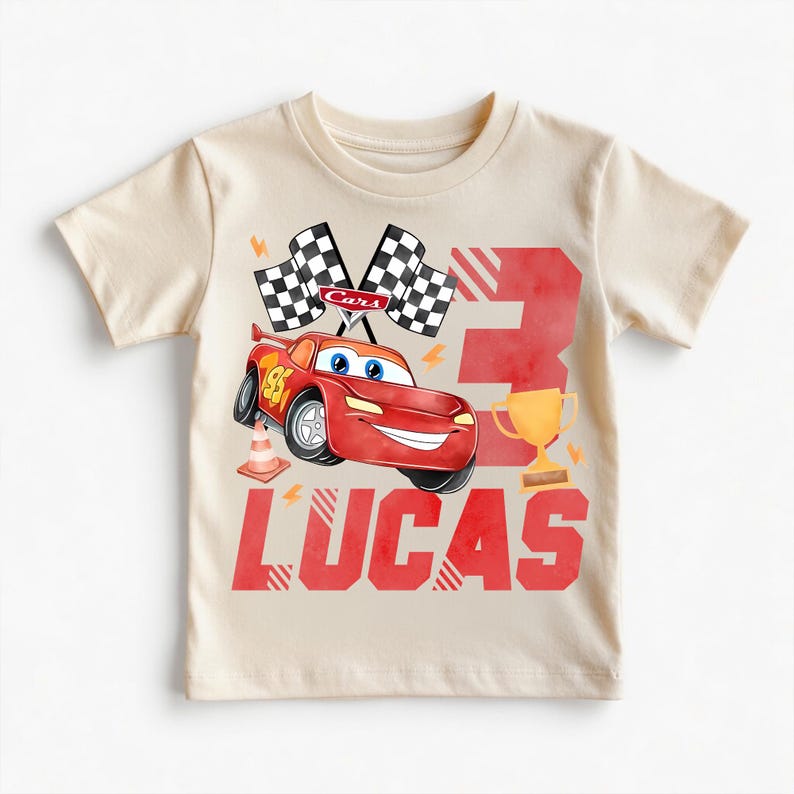 Puede incluir: Camiseta beige de manga corta con un dise&ntilde;o de coche de carreras rojo, el n&uacute;mero 95 y el nombre "LUCAS". El dise&ntilde;o incluye banderas a cuadros, un trofeo y el n&uacute;mero 3. Tambi&eacute;n se ve un cono de tr&aacute;fico.