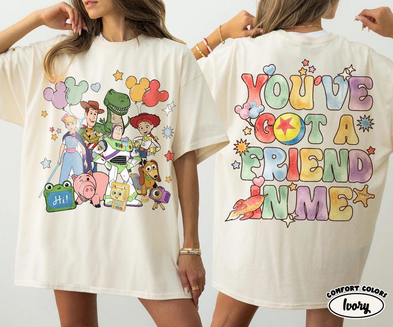 May include: An ivory t-shirt featuring a graphic print of Toy Story characters on the front and the text "YOU'VE GOT A FRIEND IN ME" in colourful, bubble-style lettering on the back, surrounded by small star, heart, and rocket illustrations.
